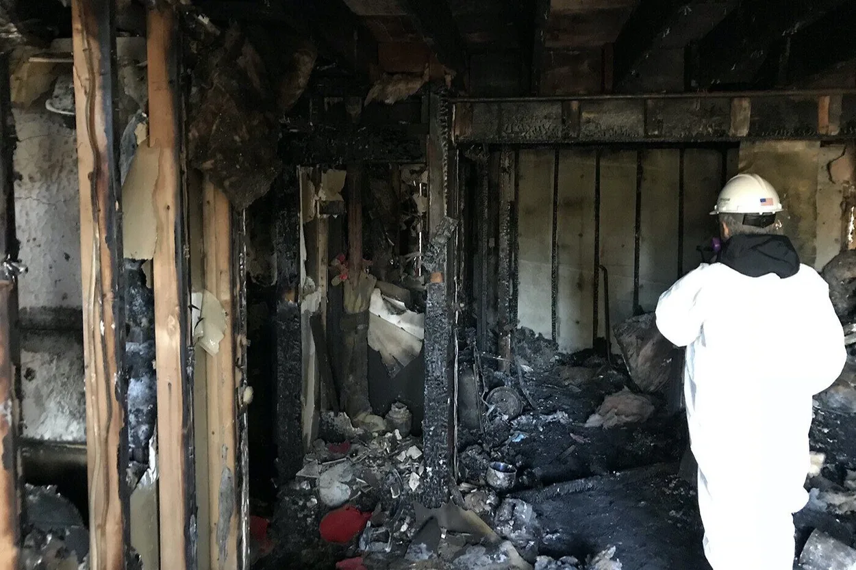 A man in a white coat stands in a burned home, assessing the damage caused by the fire.